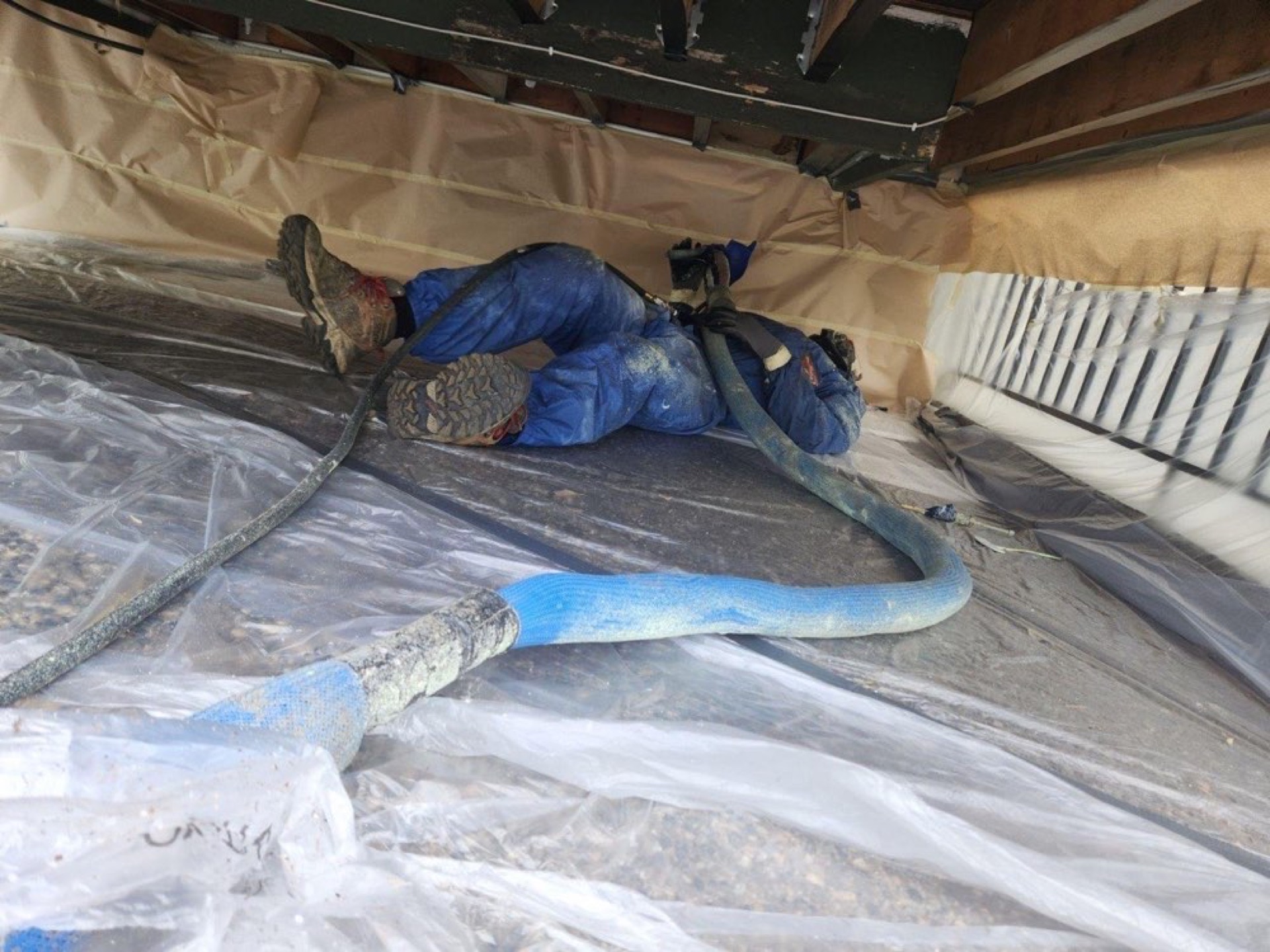 Installer working beneath floor joists in a low-clearance space with protective sheeting and a hose